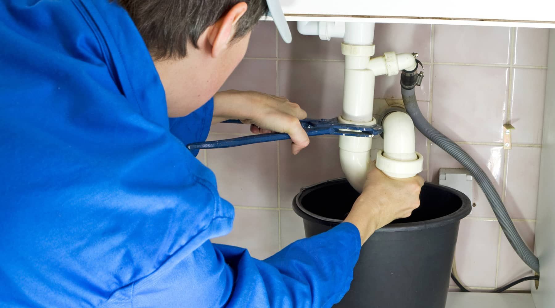 Plumber in blue uniform inspecting under-sink drain pipes with a black bucket below.