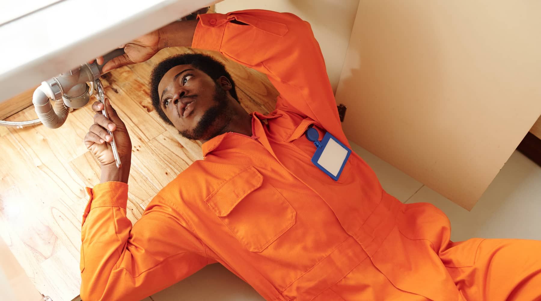 Person in orange coveralls lying on a bed, holding a tool and looking upward.