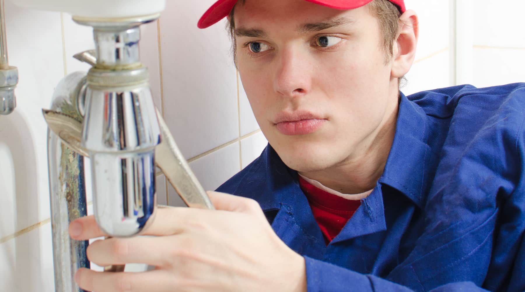 Plumber in blue overalls and red cap tightening a sink pipe under a bathroom basin