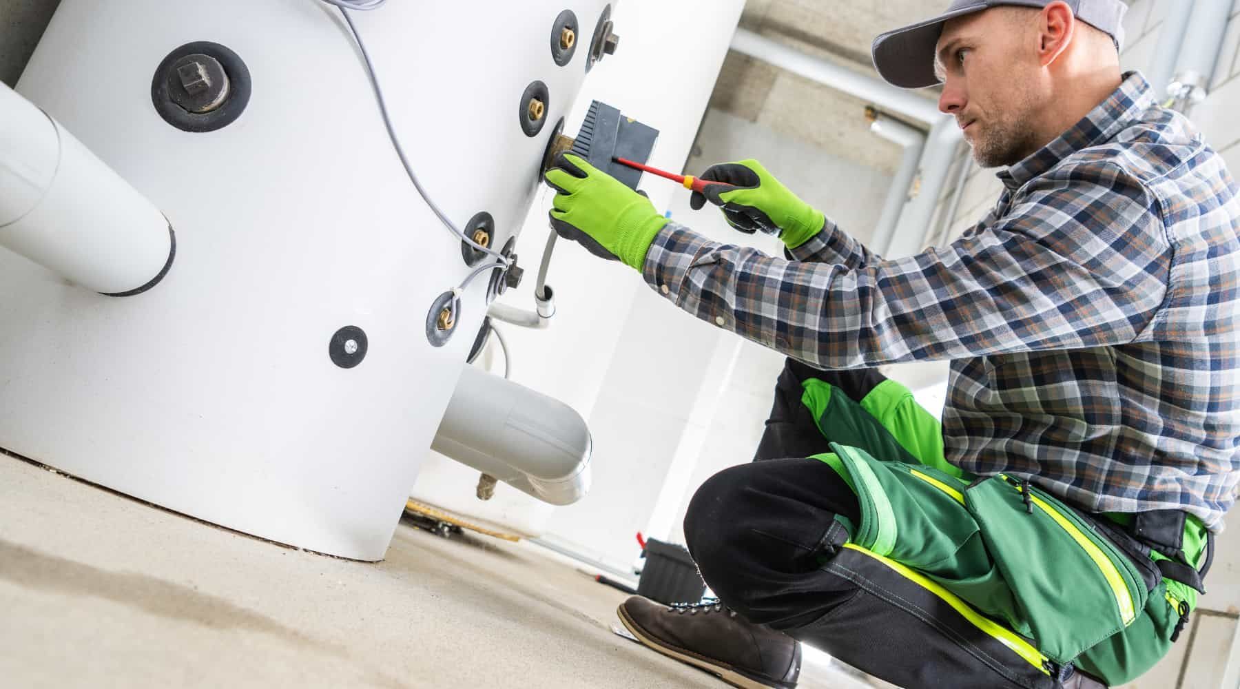 Technician in gloves and cap repairs a white appliance with a screwdriver, kneeling indoors.