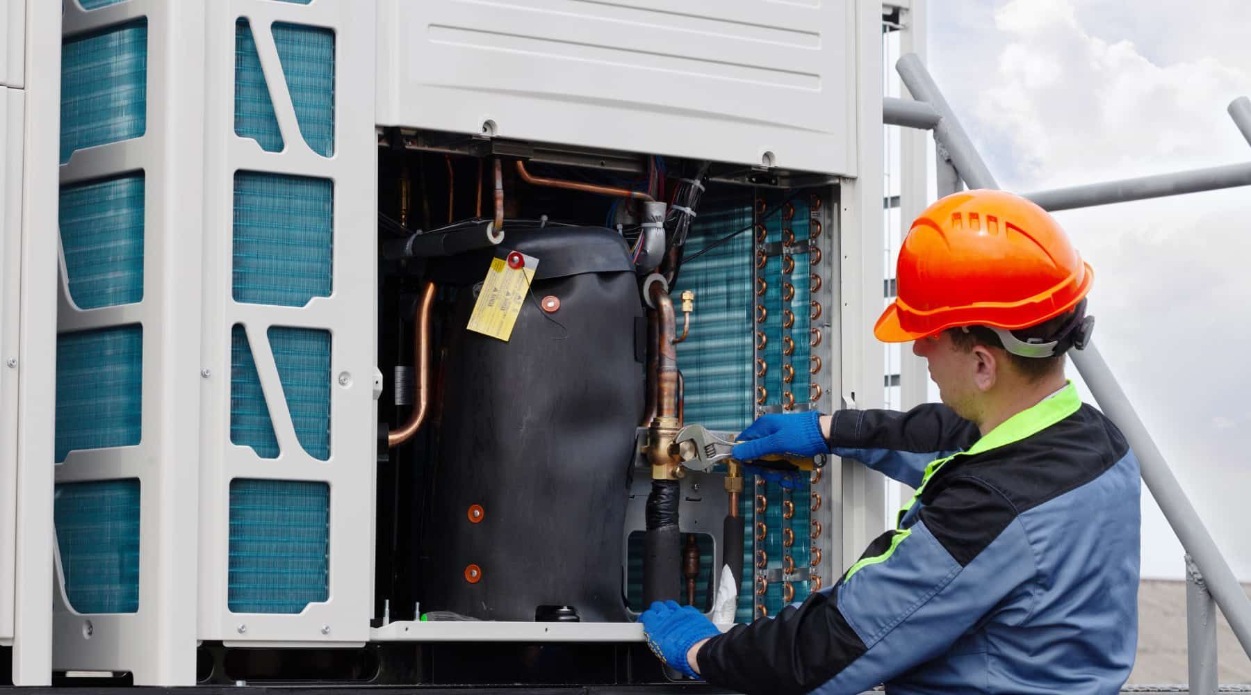 Technician in orange hard hat inspecting open outdoor HVAC unit with blue-gloved hands