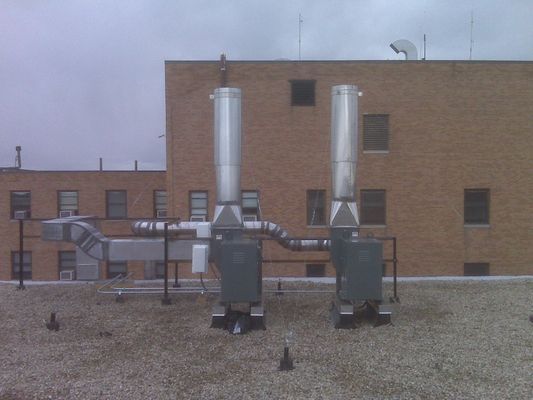 Two vertical silver exhaust stacks connected to industrial equipment on a flat rooftop in front of a brick building.
