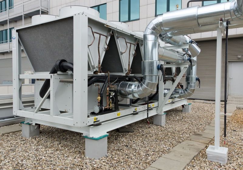 An outdoor industrial chiller unit with large silver pipes sits on a gravel roof against a building wall.
