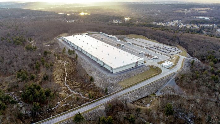 An aerial view of a large, white-roofed industrial warehouse facility surrounded by a paved parking lot and wooded area.