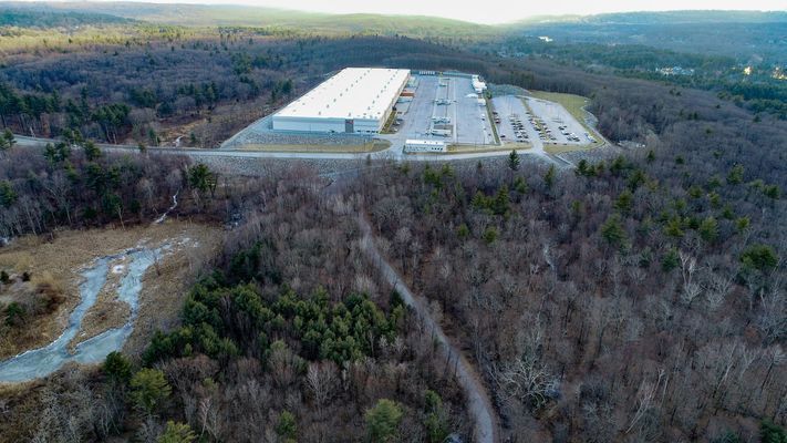 A wide aerial perspective of the warehouse facility and its associated infrastructure nestled in a hilly, forested region.