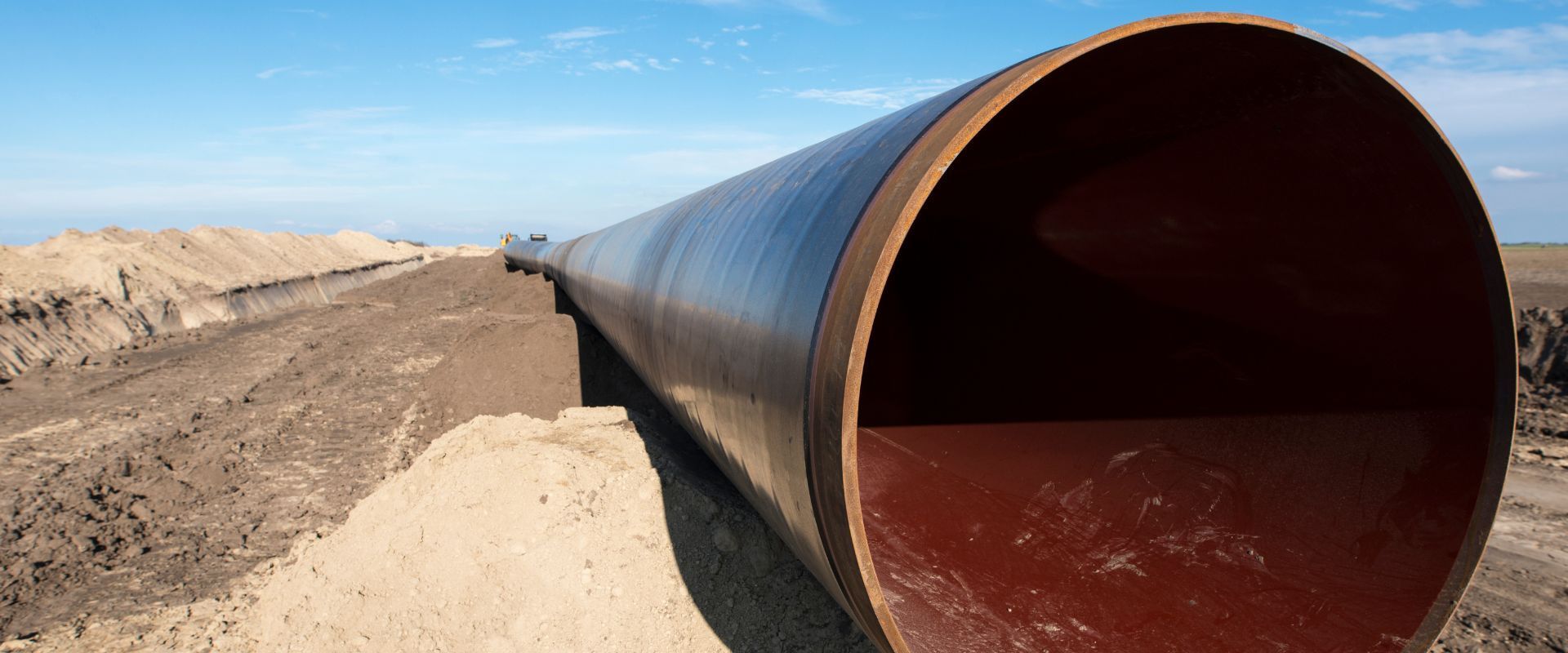 A large, dark metal pipe sits in a trench beside a dirt embankment under a clear blue sky.