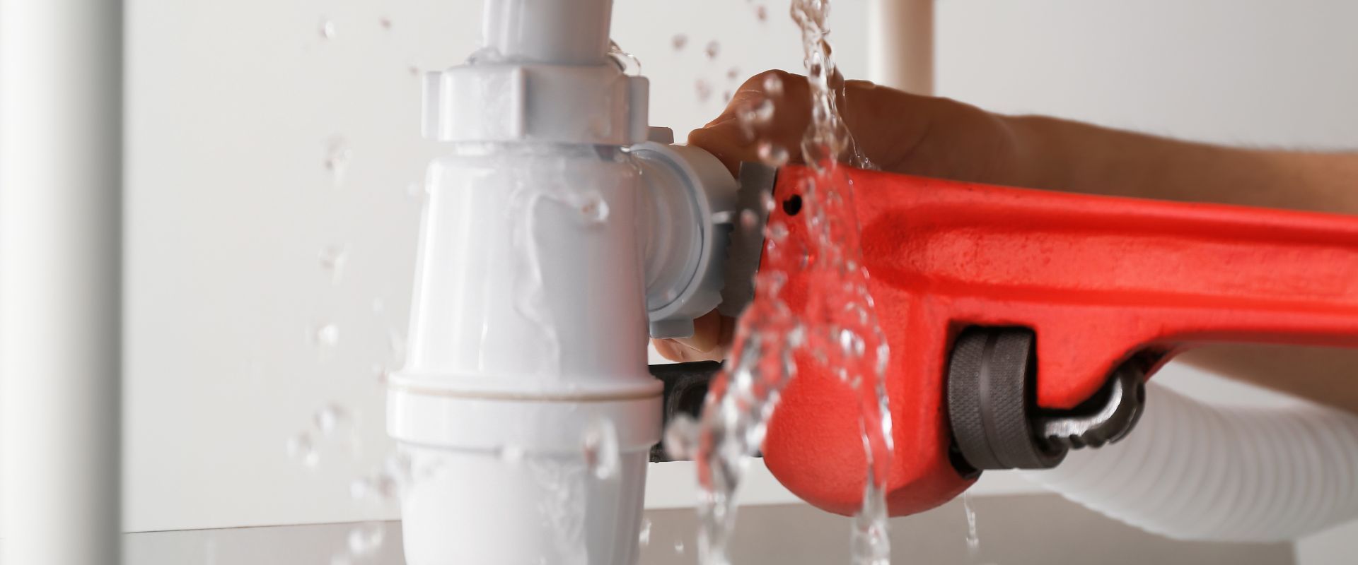 A person uses a red pipe wrench to tighten a leaking white plastic drain pipe under a sink.