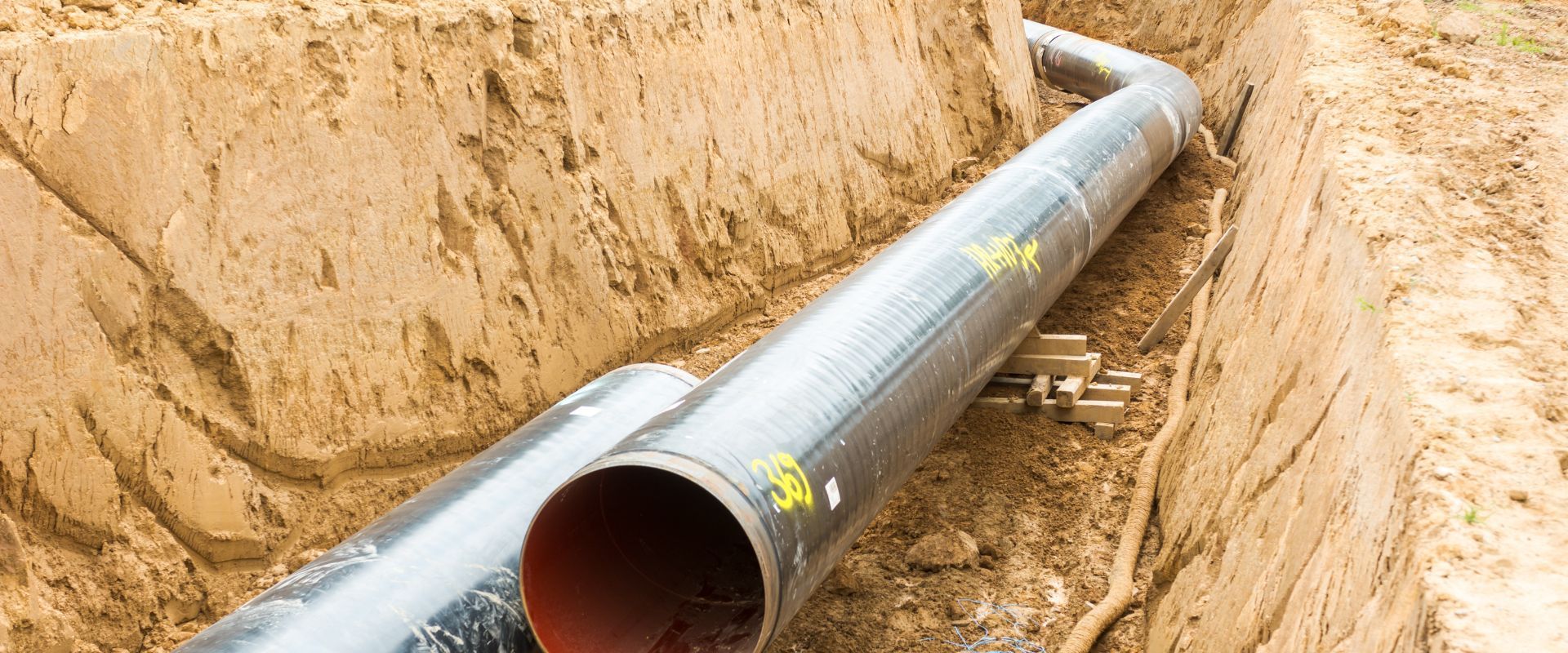 Large industrial metal pipes rest inside a deep dirt trench during an excavation or construction project.