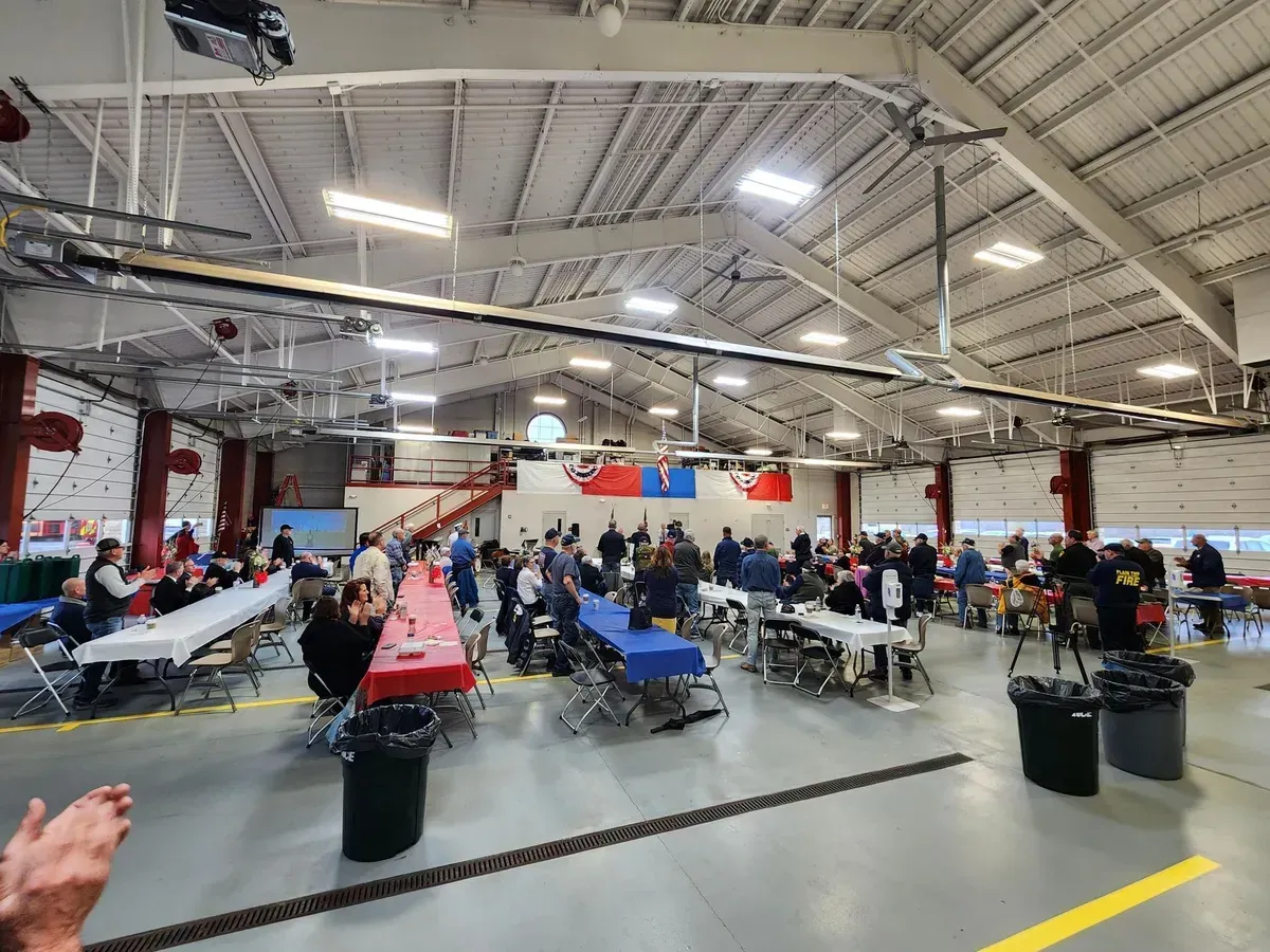 Large hangar-style room with rows of tables, people seated, and aircraft under bright overhead lights.