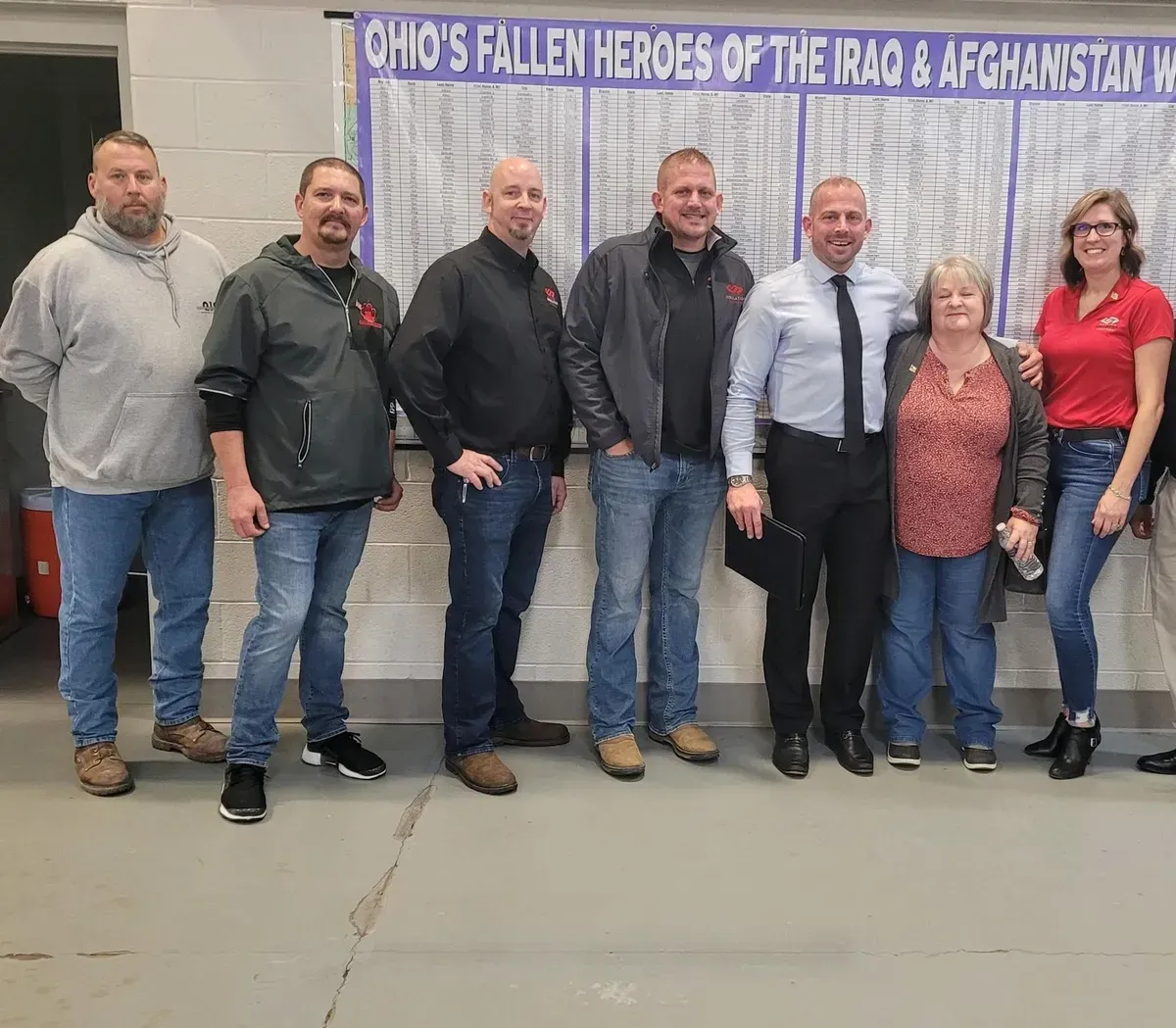 Seven adults posing indoors in front of a banner, standing shoulder to shoulder and smiling.