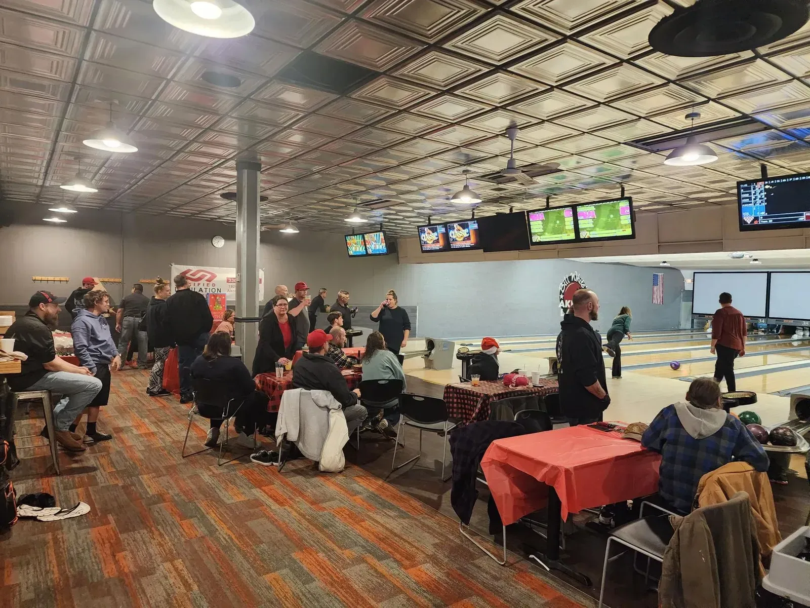 Bowling alley lounge with people seated at tables and lanes, TVs overhead, and checkered carpet.