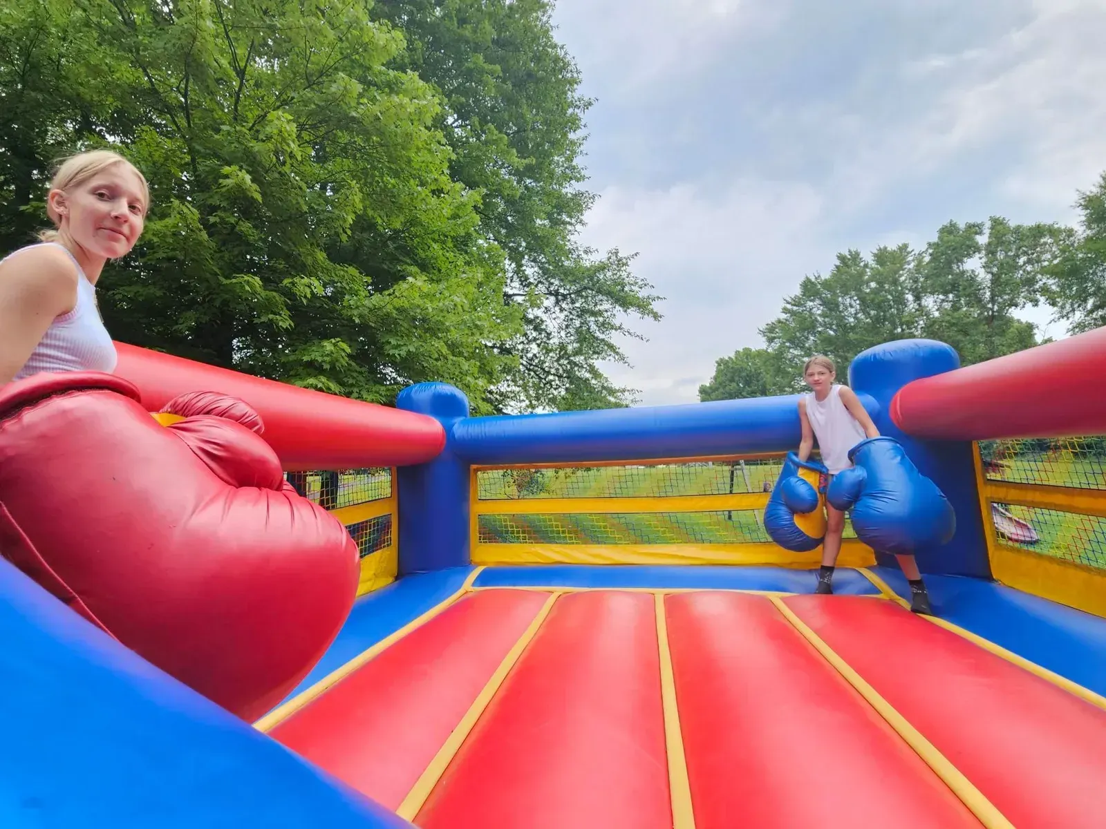 Two children on a colorful inflatable bounce house outdoors, smiling over red and blue padded walls.
