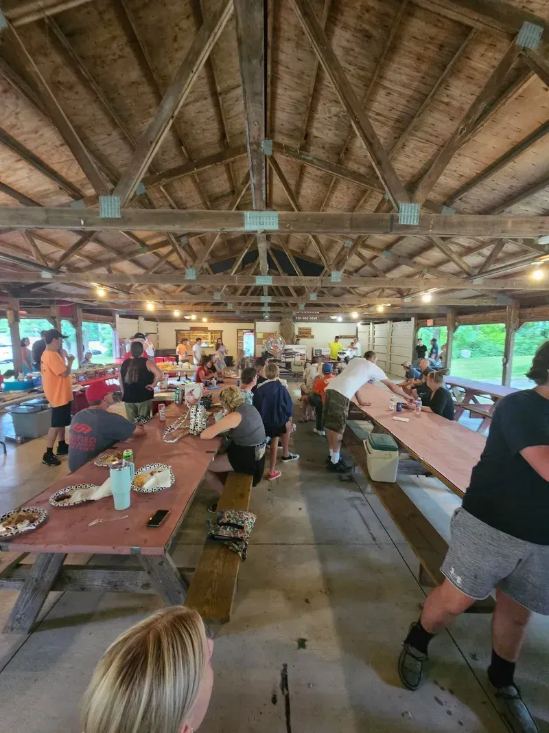 People seated at long picnic tables inside a rustic pavilion with a high wooden roof.