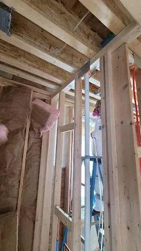 Framed interior under construction with exposed ceiling joists, pink insulation, and a ladder.