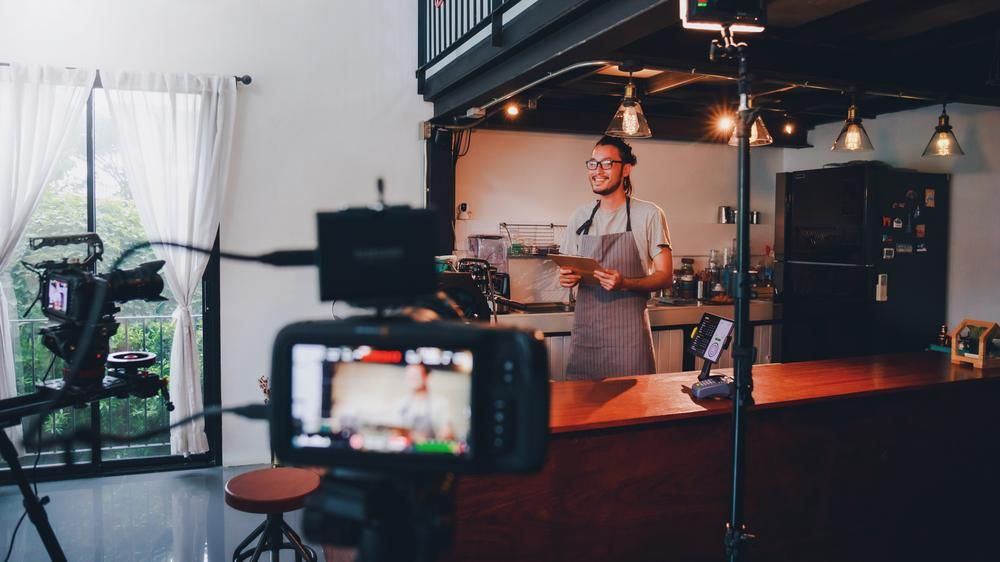 A Man in an Apron is Standing in Front of a Camera in a Kitchen — Gem Socials In Coffs Harbour, NSW