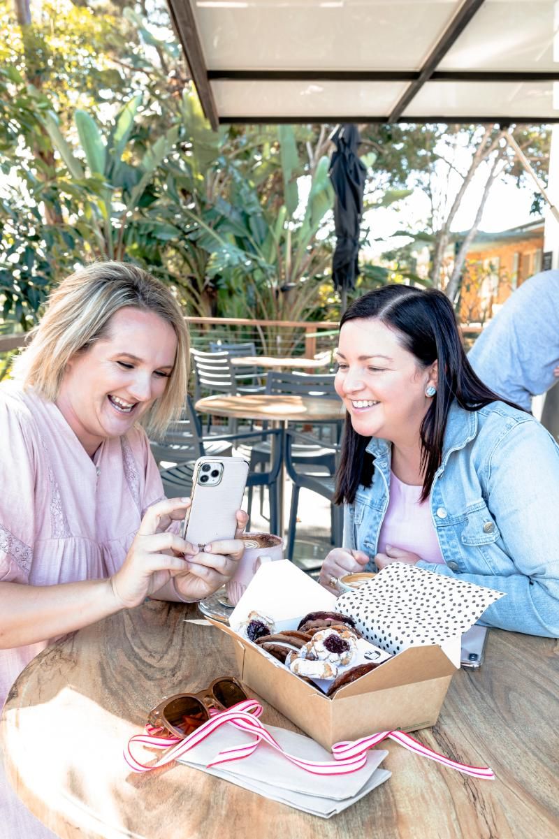 Two Women Are Sitting at a Table Looking at a Cell Phone — Gem Socials In Port Macquarie, NSW