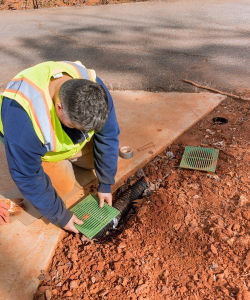 A man in a yellow vest is working on a drainage next to a sidewalk.