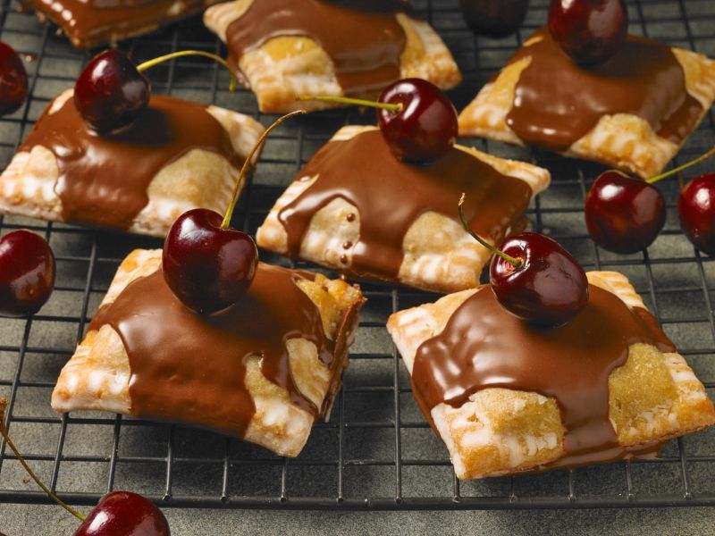 Chocolate covered pastries with cherries on top on a cooling rack