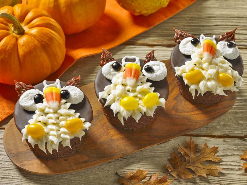 Three owl cupcakes with candy corn on them are on a wooden cutting board.