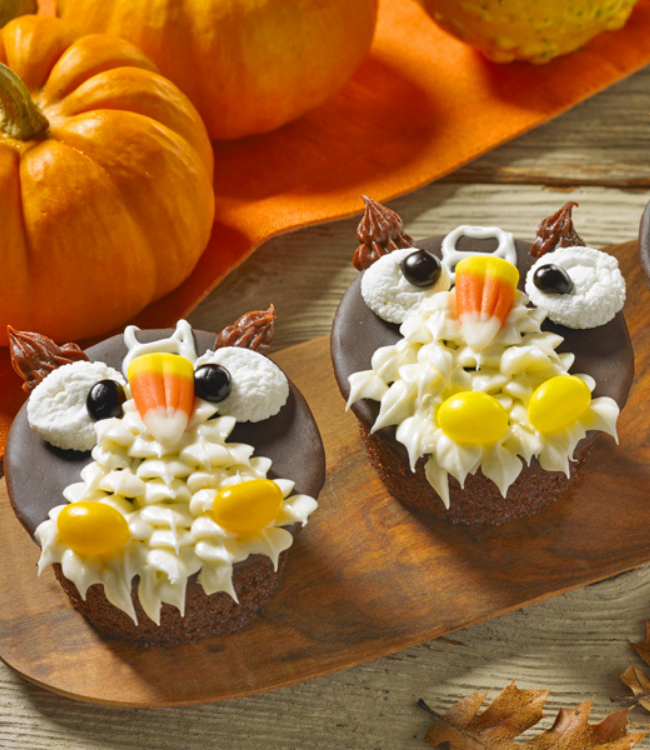 Three owl cupcakes with candy corn on them are on a wooden cutting board.