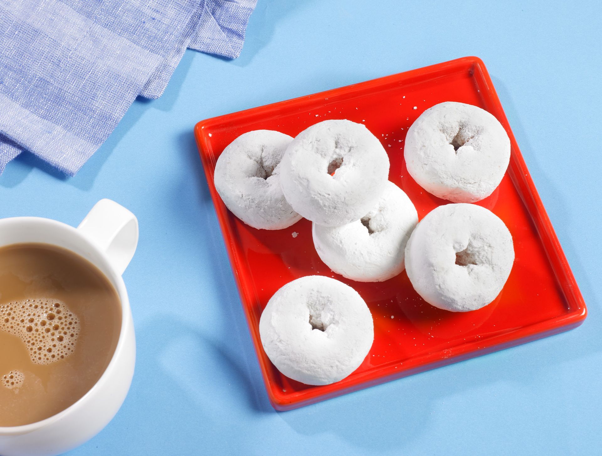 a group of powdered mini donuts on a red plate sitting on top of a blue background with a cup of coffee