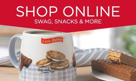 A mug with a picture of cookies on it is on a table next to a plate of food. Text is 