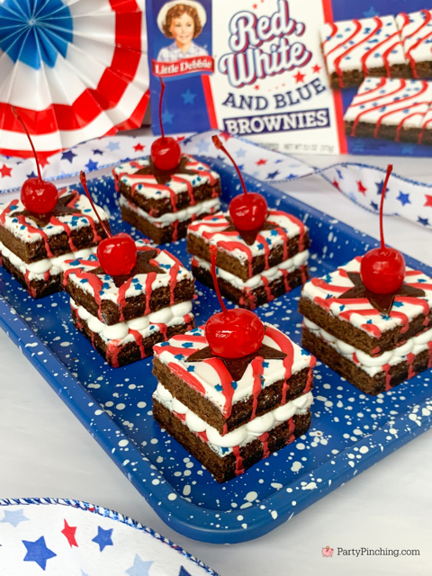 Close-up of festive brownie bites topped with whipped cream and cherries, arranged on a blue speckled tray with red and white decorations.