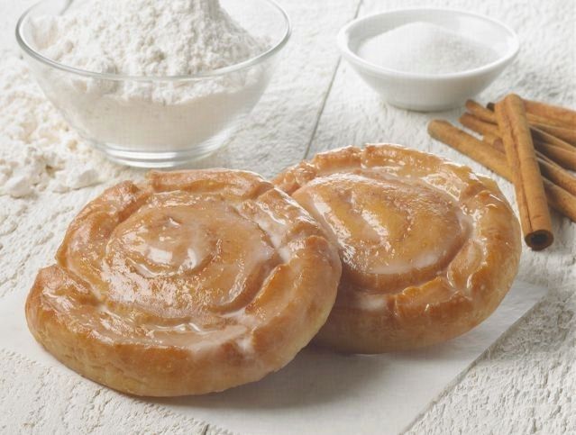 Two cinnamon rolls are sitting on a table next to a bowl of flour and cinnamon sticks.