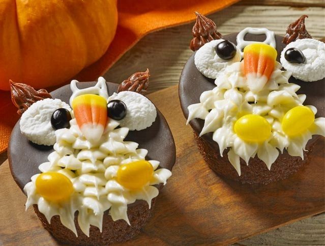 Three owl cupcakes with candy corn on them are on a wooden cutting board.