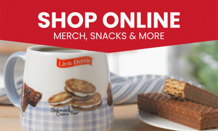 A mug with a picture of cookies on it is on a table next to a plate of food. Text is 