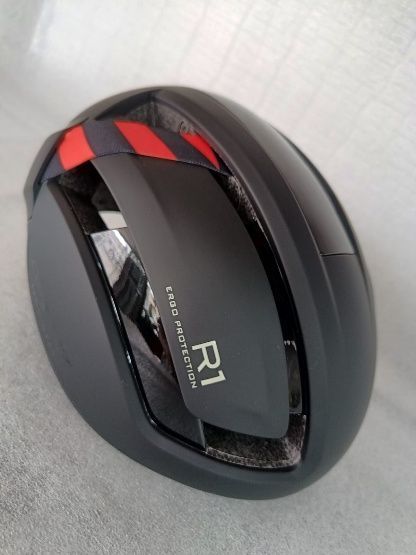 Dispositivo R1 ovalado de color negro con detalles en rojo, mostrado en ángulo sobre una superficie clara.
