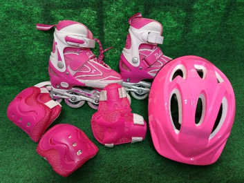 Patines de ruedas rosas, casco, rodilleras y muñequeras dispuestos sobre césped verde.