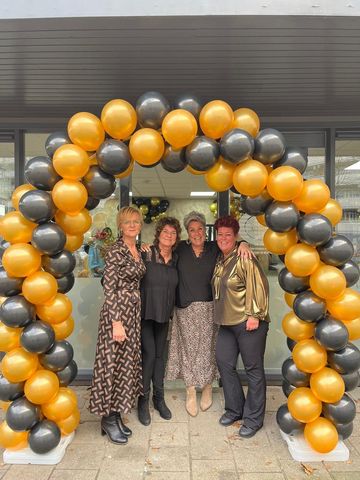 Vier mensen staan onder een ballonnenboog van gouden en zwarte ballonnen voor een gebouw.
