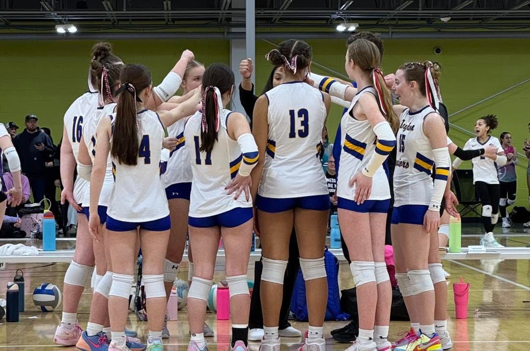 Volleyball team huddles on court in white and blue uniforms, numbers visible, in an indoor gym.