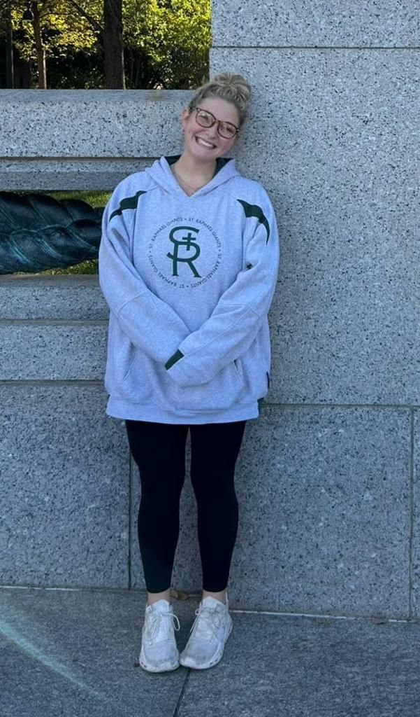 Person smiling in a gray sweatshirt, black leggings, and white shoes, standing against a stone wall.