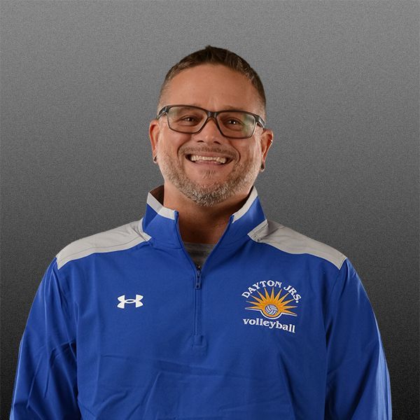 Smiling man in blue athletic jacket with Dayton volleyball logo, standing against a gray background