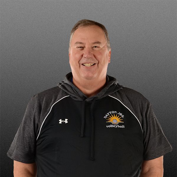 Smiling man in a dark hoodie with a volleyball logo, standing against a gray background