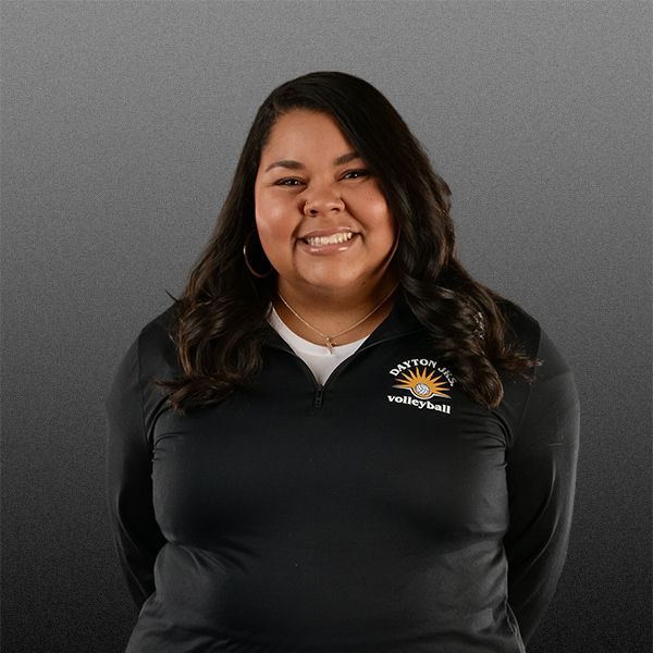 Smiling person in a black polo shirt with a sun logo, standing against a gray background.