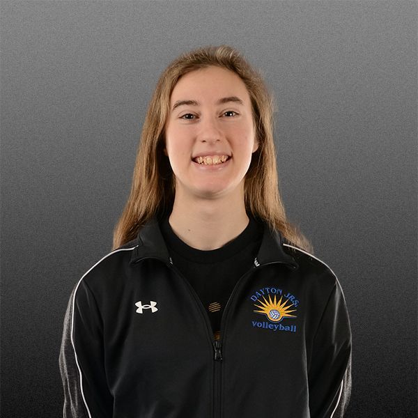 Person in a black jacket with a sports logo, smiling against a gray background.