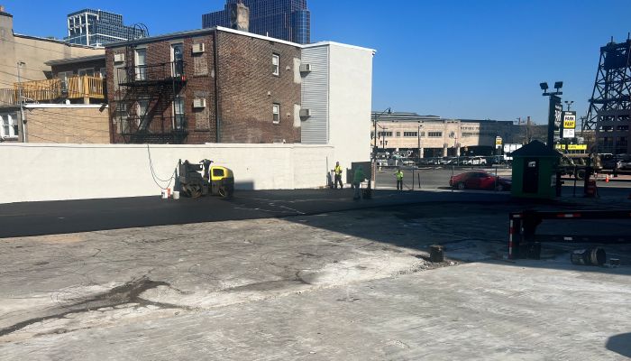 Construction workers paving a parking lot near brick buildings on a sunny day.