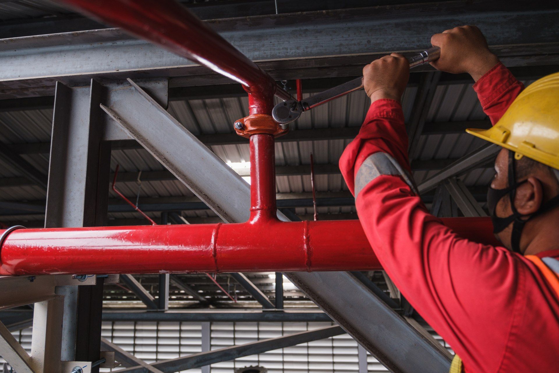 A person in safety gear tightens a red pipe with a wrench indoors, near metal beams.