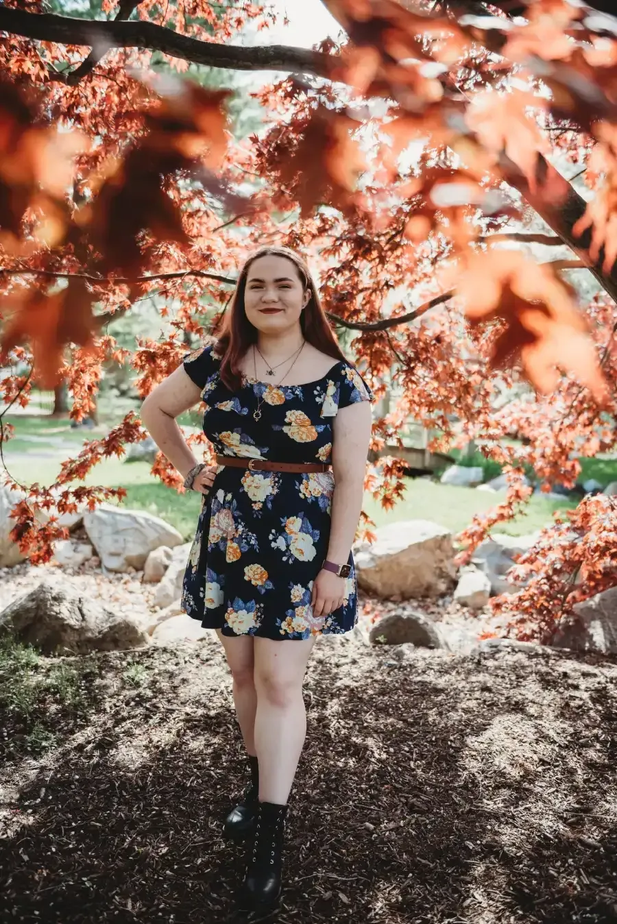 Woman standing under pink flowering tree branches in a garden, wearing a navy floral dress and black boots.