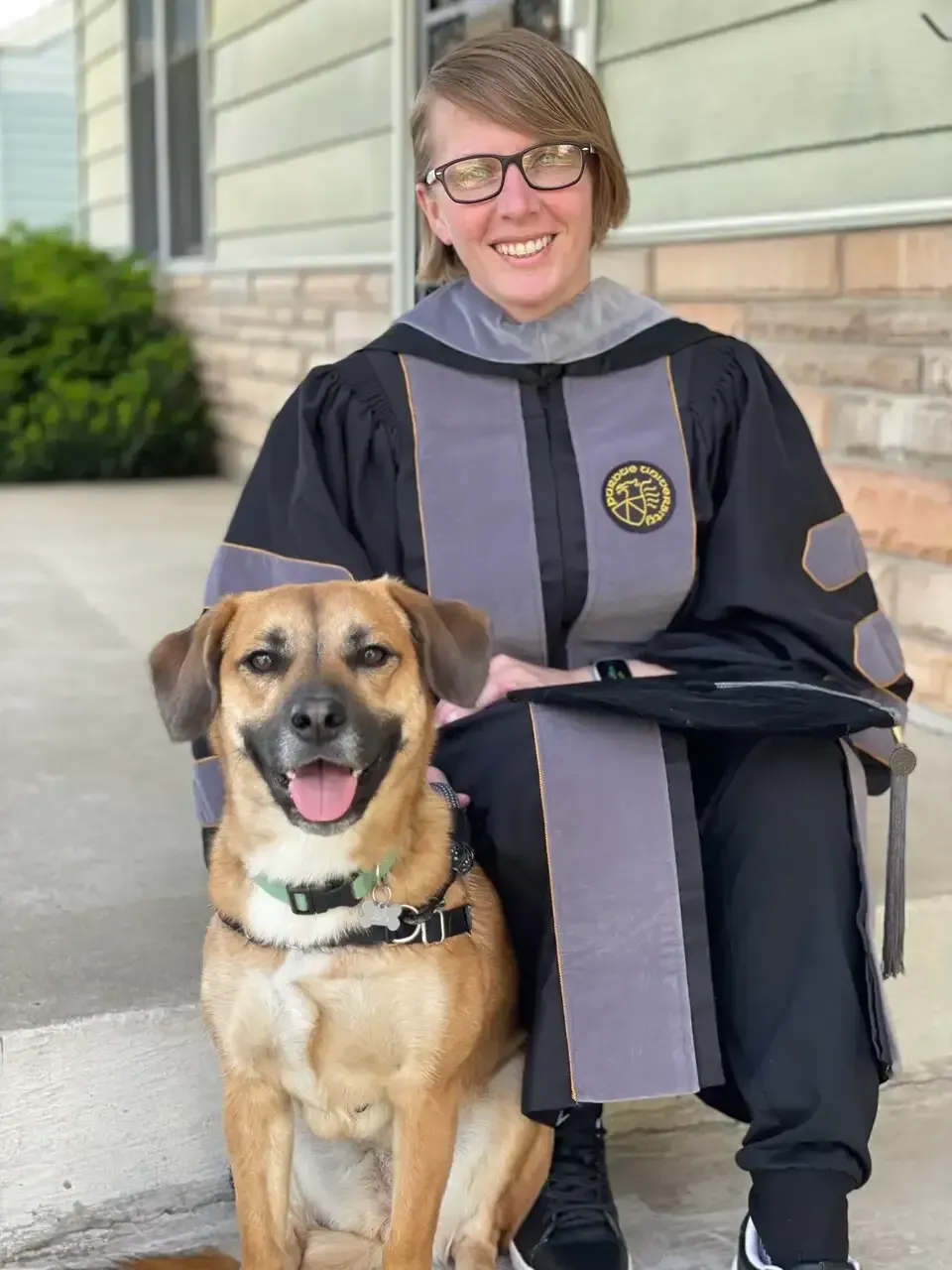 Person in graduation robe kneels beside a smiling dog on a porch