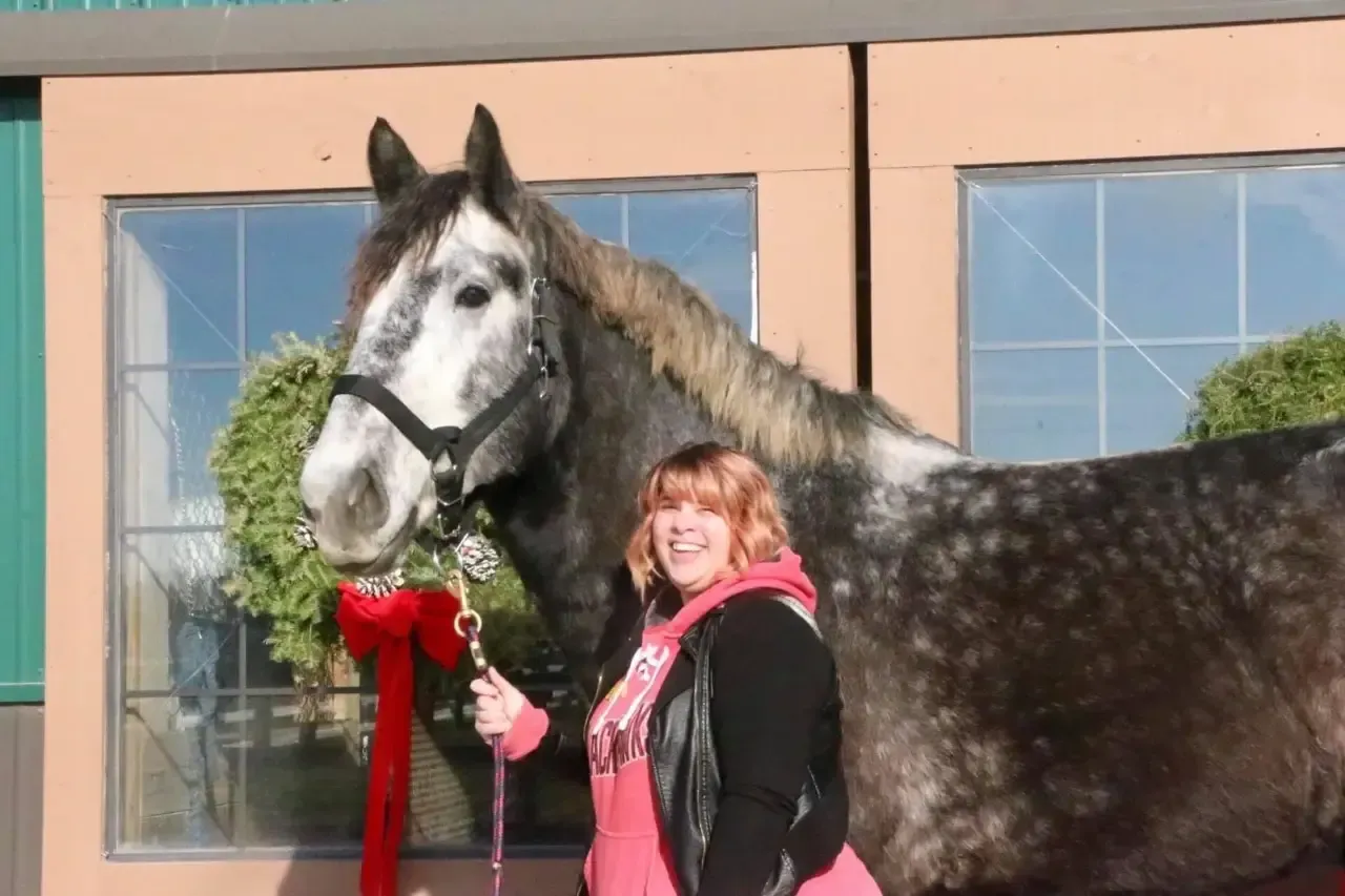 Woman stands beside a gray horse with a red ribbon in front of a building.
