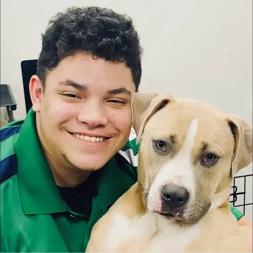 Smiling person in a green shirt holding a tan-and-white dog indoors