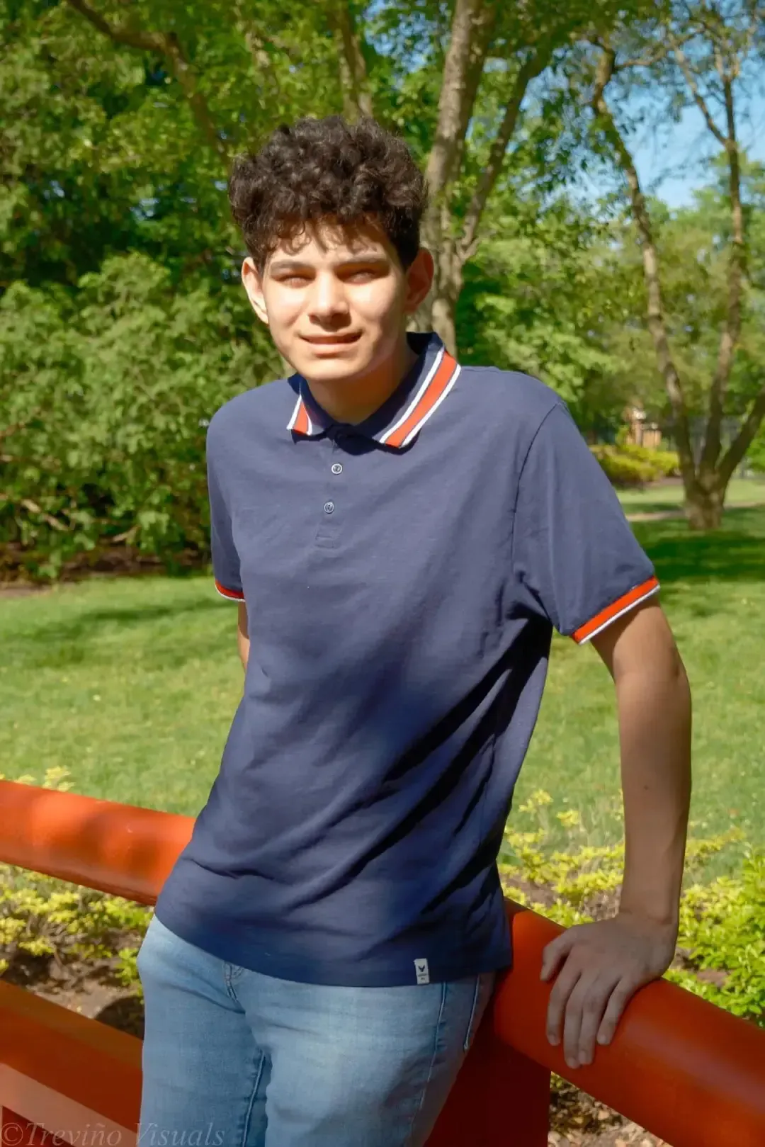 Person leaning on an orange rail in a sunny park, wearing a navy polo and jeans.