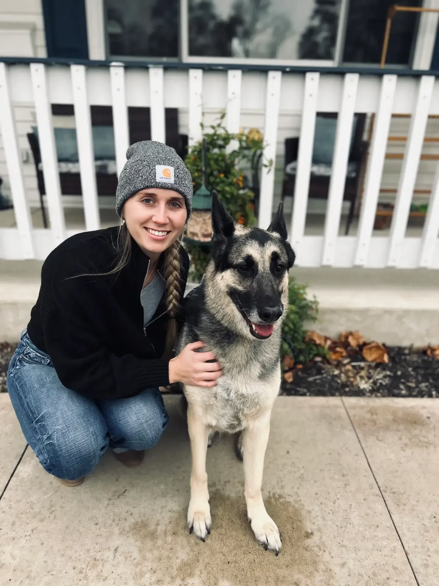Person kneels beside a smiling German Shepherd on a sidewalk by a white fence.