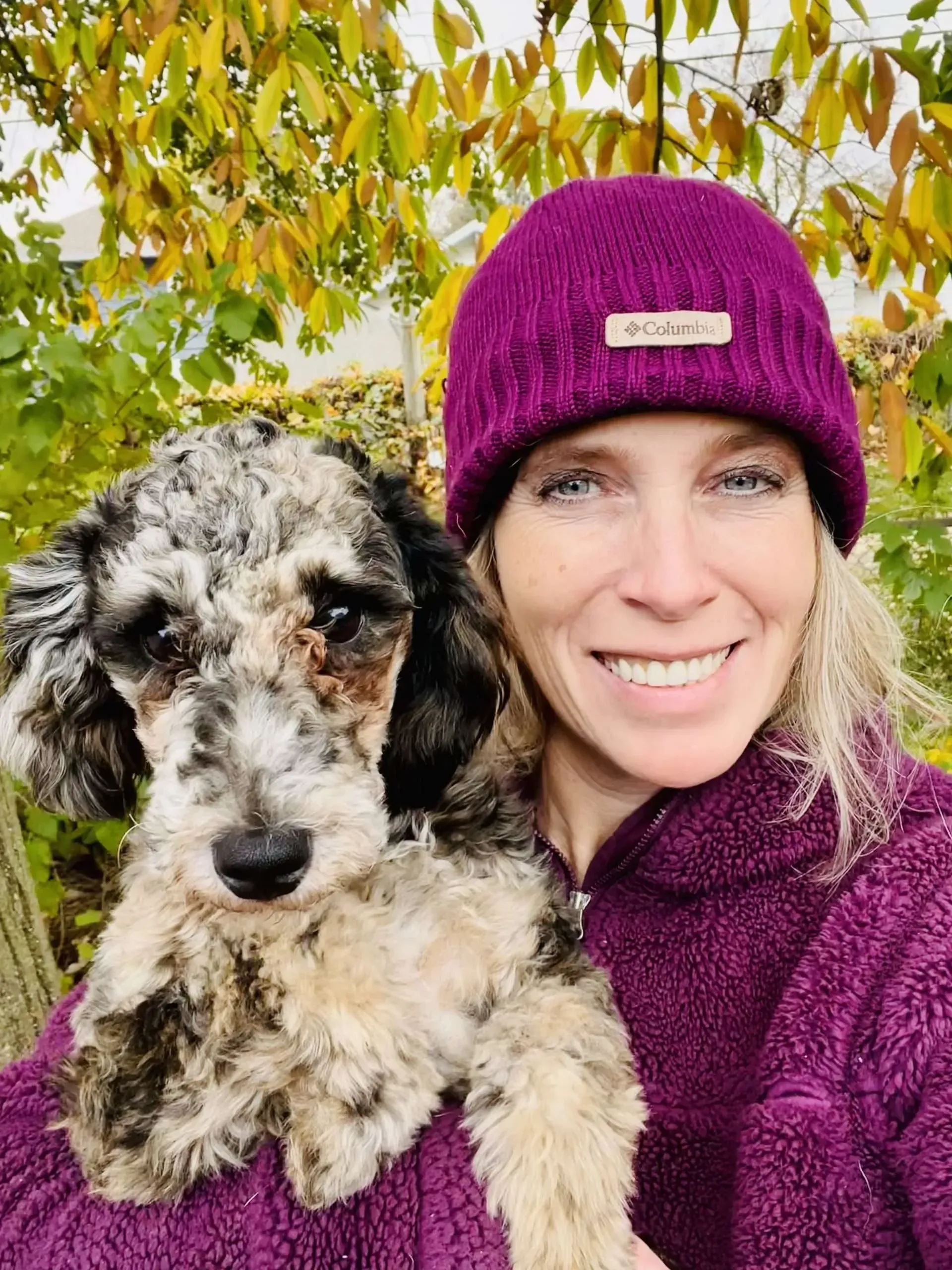 Woman in a magenta beanie holding a black-and-white dog outdoors among autumn leaves