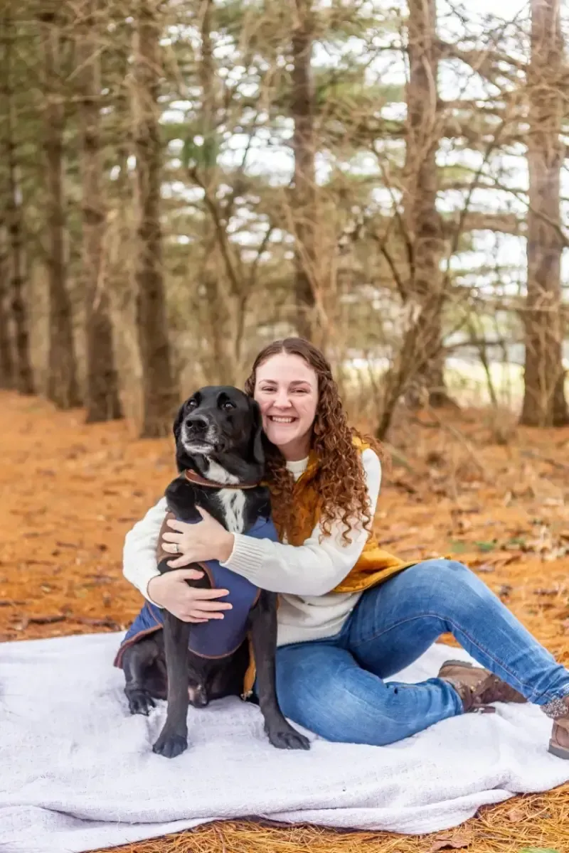 Woman sitting on a blanket in a forest, hugging a black dog in a sweater