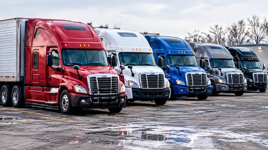 Four semi trucks in red, white, blue, and black lined up on a wet lot under an overcast sky